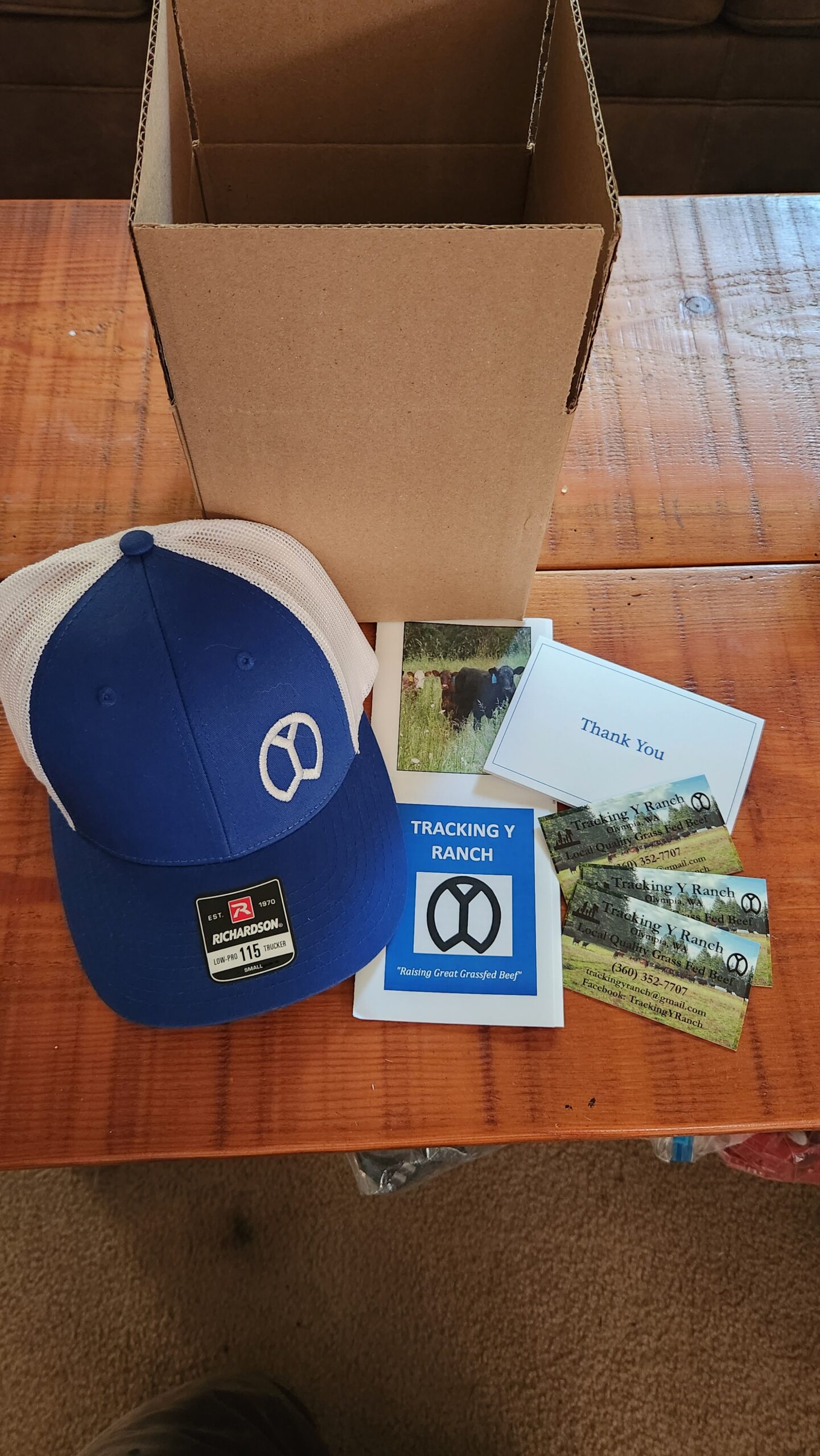 Blue and white cap from Tracking Y Ranch, greeting cards, business cards, and a thank you note arranged on a wooden table next to an empty cardboard box. The items promote grass-fed beef and local farming.