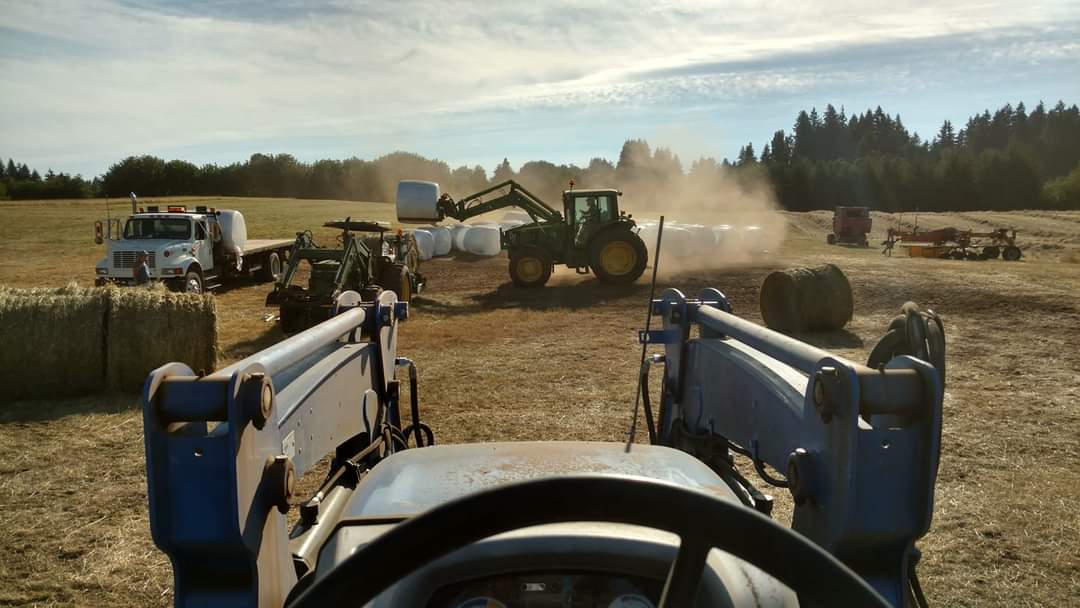 View from the driver's seat of a tractor on a farm, showcasing various farming equipment including a green tractor lifting hay bales, a white truck, and additional machinery in a dusty field under a clear blue sky.
